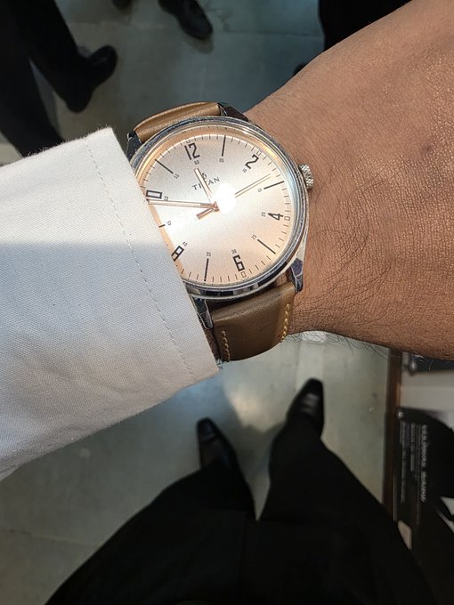 A close-up of a wrist wearing a 'TRIAN' watch with a brown leather strap and silver dial, with the wearer dressed in a white shirt.