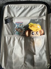 An open suitcase with a light-colored interior, containing a grey mesh zippered pocket holding two small plush toys—one resembling a character with a colorful headdress and another orange and white character.