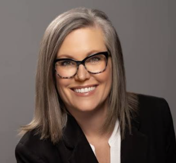A professional headshot of a smiling woman with long gray hair and tortoiseshell glasses, wearing a black blazer over a white top. The image appears to be a stock-style portrait, likely used to illustrate Katie Hobbs, the Arizona governor mentioned in the post who has vetoed Bitcoin-related legislation three times.