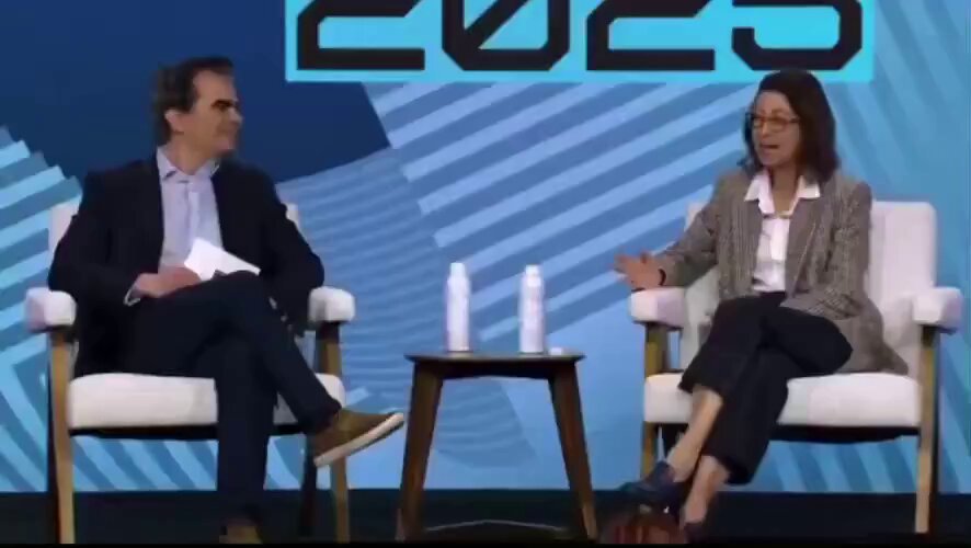 The image features a man and woman sitting across from each other at a table, engaged in conversation. They are both wearing suits and glasses, suggesting they might be professionals or attending a formal event. The table between them has two bottles on it, possibly related to their discussion about Bitcoin as the focal point of the post.

The background shows a blue wall with white stripes, which could be part of an event setup for the conversation. There is also a chair visible in the scene, indicating that they are seated at a table or a similar surface during their discussion. The image does not contain any text related to Bitcoin or its value, but rather focuses on the man and woman's interaction with each other.