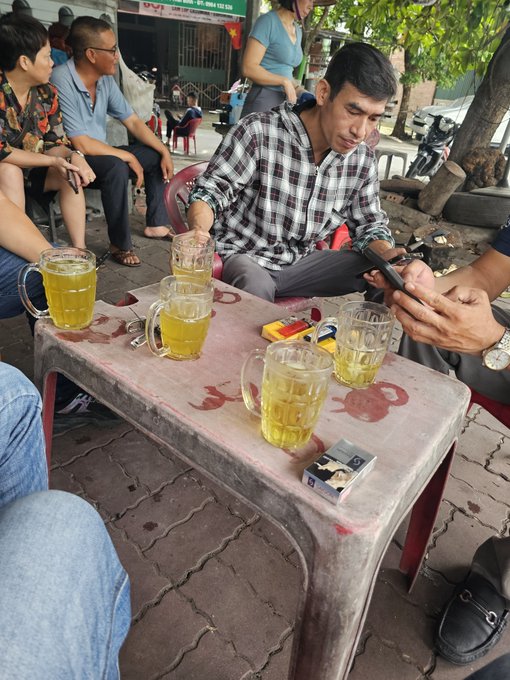 A group of people gather around a small stone table at an outdoor café, with several glass mugs of tea, a pack of cigarettes, and one person looking at their phone.