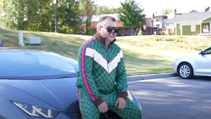 A man wearing a green Gucci tracksuit with white stripes sits on the hood of a blue car, captioned to joke about being the "whitest" among a group of "white bulls," referencing the white chevron design on his tracksuit as a play on the term.