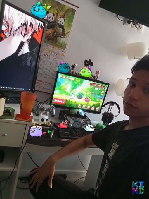 A person sits at a gaming setup with two monitors displaying anime art and a game called 'RABUK K', surrounded by various cartoon character stickers and decorations, with 'KI ND' watermarked in the corner.