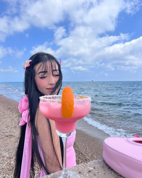A young woman with long dark hair wearing a pink outfit and flower hair clips at a beach, posing with a pink margarita cocktail garnished with an orange slice against a blue ocean and cloudy sky backdrop.
