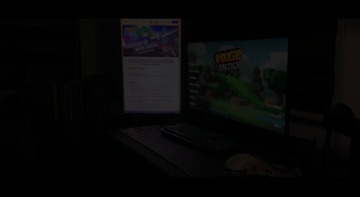The image shows a computer monitor displaying a video game called "POV" (Post Office voxie), with an accompanying website or blog post about it, which reads "@VoxieTactics". The background appears to be dark, but there is also some light coming from behind the screen. There are books on either side of the image and a mouse near the bottom right corner.