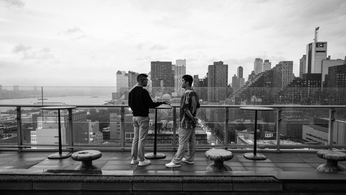 Two men in business casual attire converse on a rooftop terrace overlooking a city skyline with waterfront views, fitting the professional networking atmosphere of the Tokenize NYC conference where Newton mainnet beta was launched.