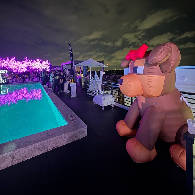 A nighttime rooftop party scene featuring an illuminated pool, pink-lit trees, and a large inflatable brown bear with a red bow, with people gathered in the background.