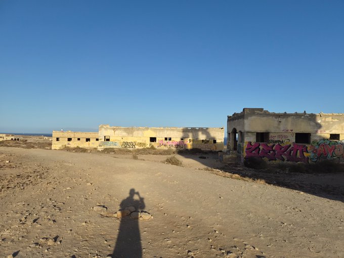 A photograph of abandoned, graffitied concrete buildings in a desert landscape under a clear blue sky, with the photographer's shadow visible in the foreground.