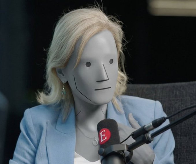 A blonde woman wearing a light blue blazer being interviewed at a microphone with an 'E.' logo, while wearing a blank white featureless mask over her face.