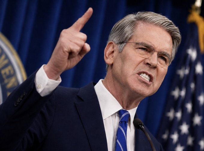 A man in a dark suit and blue tie speaks emphatically at a podium, pointing upward with his index finger against a backdrop of American flags. The image, accompanying news of U.S. Treasury Secretary Bessent's announcement regarding upcoming bank sanctions, captures a moment of decisive political communication.