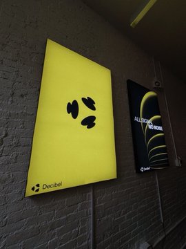 The image features two posters hanging side by side against a brick wall, both with yellow backgrounds and black text. The first poster has a picture of three circles, while the second poster displays an advertisement for "All-in-One Markets". Both posters are illuminated, making them stand out prominently on the wall.