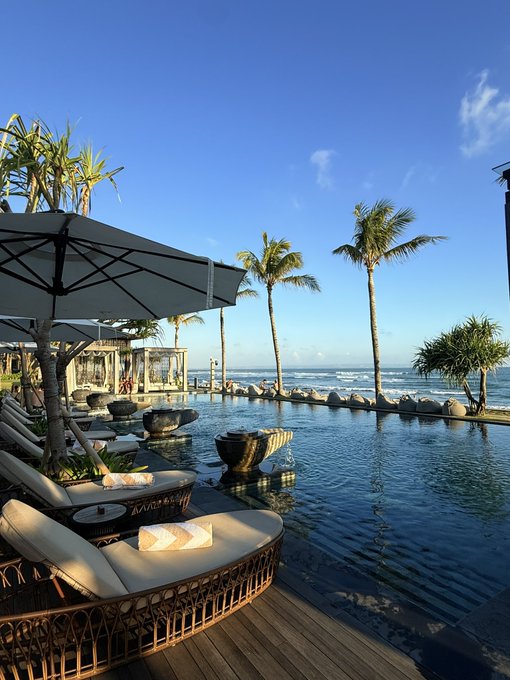 A serene oceanfront resort features palm trees swaying beside an infinity pool, with lounge chairs and umbrellas on a wooden deck overlooking the sea. The image pairs with the post's message about staying grounded on the journey toward a 200k Bitcoin goal, reflecting the calm, scenic perspective of treating market dips as passing views rather than setbacks.