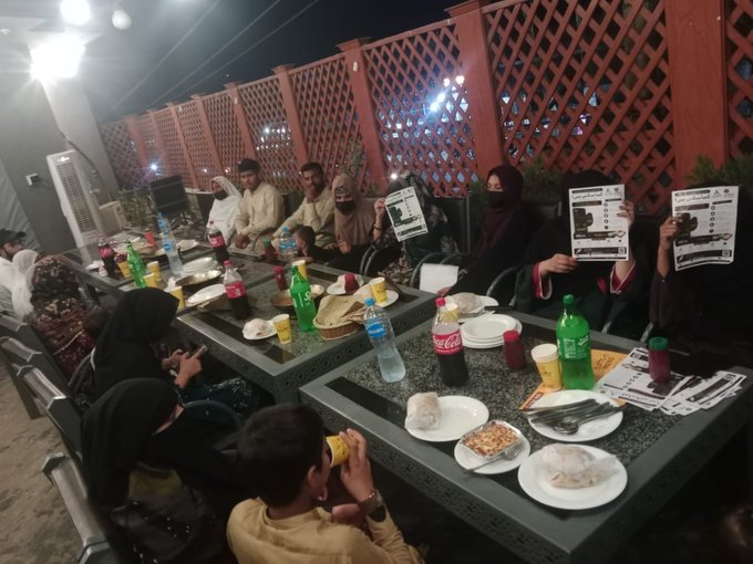 A group of men and women gather around tables laden with Coca-Cola bottles, water, and plates of food at an outdoor evening meetup, with two women on the right proudly holding up AILYTIS-branded promotional flyers, illustrating LV1 Leader Saith Sahib's offline gathering in Pakistan to introduce AILYTIS and ODBIT while fostering local team connections.