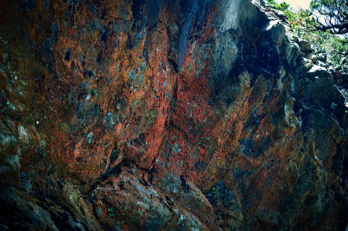 A close-up photograph of a rocky cliff face displaying streaks of red, orange, and dark mineral coloration.