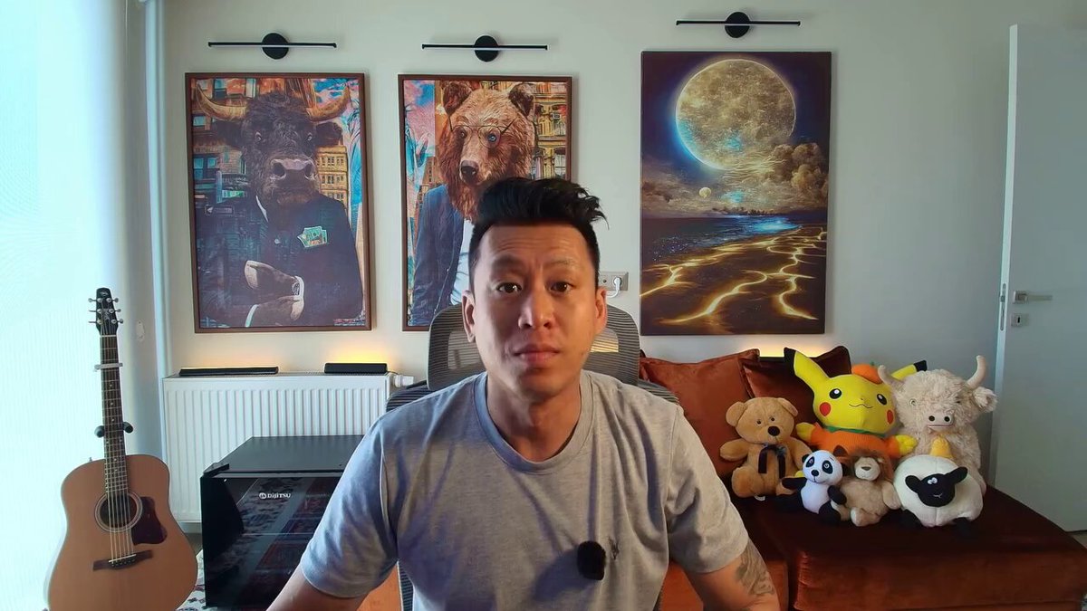 The image shows a man sitting in front of a couch with several stuffed animals around him. The man has a serious expression as he looks directly at the camera. There are also two paintings hanging above the couch, one depicting a bear and another featuring a cartoon character. A guitar is placed next to the couch on the left side of the image.