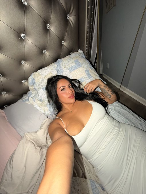 A woman with long dark hair takes a selfie in a white dress while lying on a bed with a tufted silver headboard.