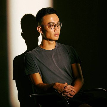 A man with glasses, wearing a gray t-shirt, sits with his hands clasped as warm light casts a dramatic shadow on the wall behind him. The contemplative pose aligns with the accompanying quote about Hyperliquid CEO Jeff Yan's focus on originality and pioneering new ideas.