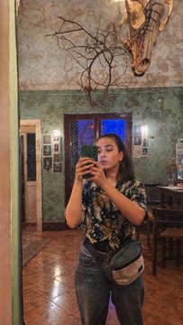 A mirror selfie of a young woman taking a photo with her phone, wearing a patterned short-sleeve shirt and jeans with a green crossbody bag in a uniquely decorated room with patterned walls and a hanging animal skull.