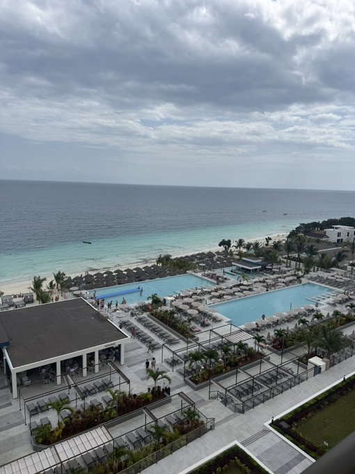 An elevated view of a tropical beachfront resort featuring two large swimming pools, rows of lounge chairs, palm trees, white cabanas, and a turquoise ocean under a cloudy sky.