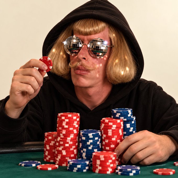 A man wearing a black hoodie, a blonde bob wig, a mustache, and disco ball sunglasses holds a red poker chip above stacks of chips on a green table.