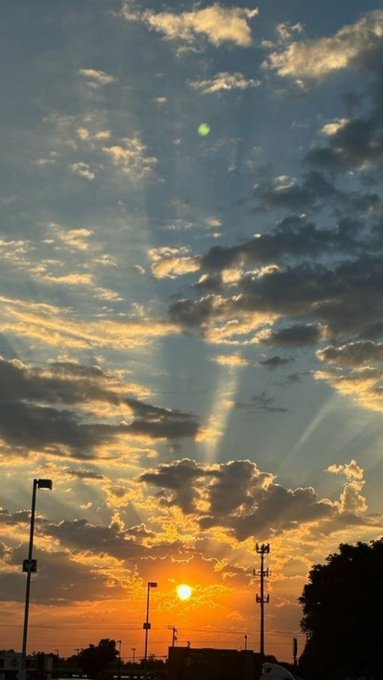 A vibrant sunset with rays radiating through scattered clouds over a cityscape, with light poles and rooftops silhouetted against the horizon.