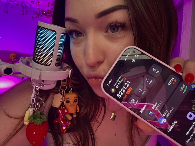 A woman with red nails holds a smartphone showing a stock portfolio app with a total of $22.21, while a white microphone with colorful charms is visible in the foreground against a purple-lit background.