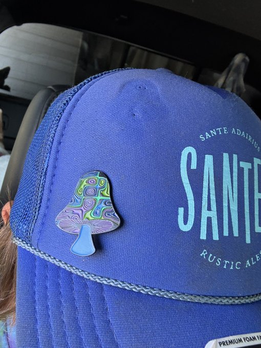A close-up of a blue trucker hat branded 'SANTE ADAIRIUS RUSTIC ALE' featuring an iridescent mushroom pin attached to the front.