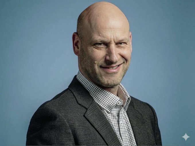 The image features a bald man wearing a suit jacket and tie, smiling at the camera with his eyes closed. The background of the photo is blue, which contrasts nicely with the man's attire. There are also some stars visible in the bottom right corner of the image. The caption provided seems to be unrelated or unrelated to the main subject matter of the image - a man wearing a suit and tie. It could possibly be an advertisement for something else, but it doesn't seem directly related to the man in the photo.