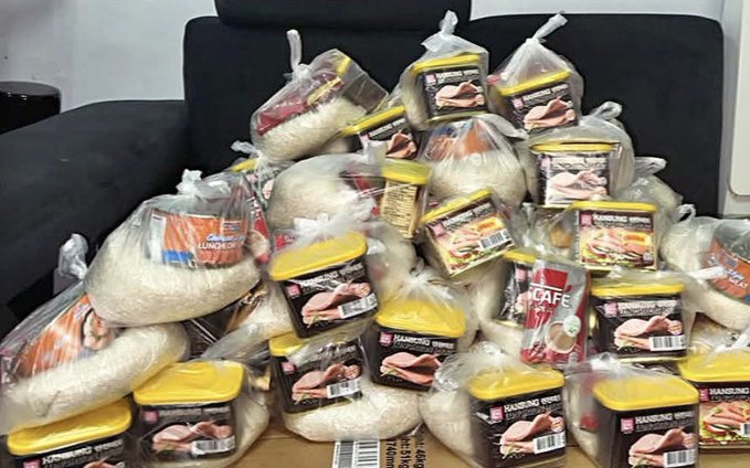 A pile of packaged food items including bags of rice and containers of sliced ham stacked on a surface in front of a black sofa.