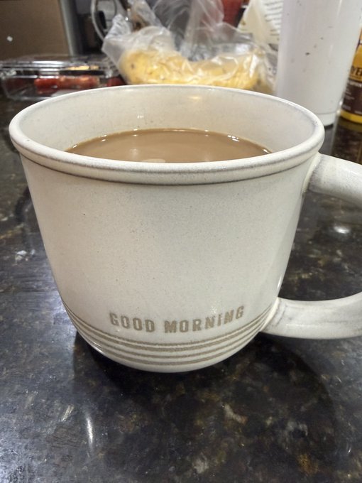 A close-up of a ceramic coffee mug filled with coffee and milk, with the text "GOOD MORNING" printed on its side, sitting on a dark countertop.