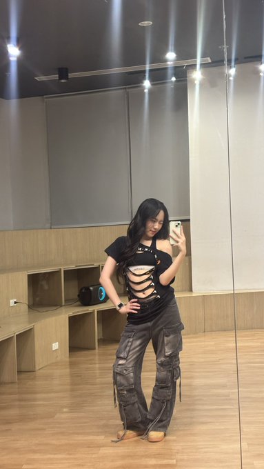 A woman takes a mirror selfie in a dance studio, wearing a black cut-out top and dark cargo pants with strap details.