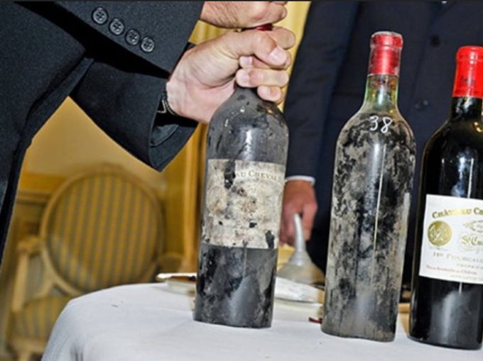 A man in a dark suit pulls a cork from an old, dusty vintage bottle of Chateau Cheval Blanc wine, with another moldy bottle labeled '38' and a third bottle visible on a white tablecloth.