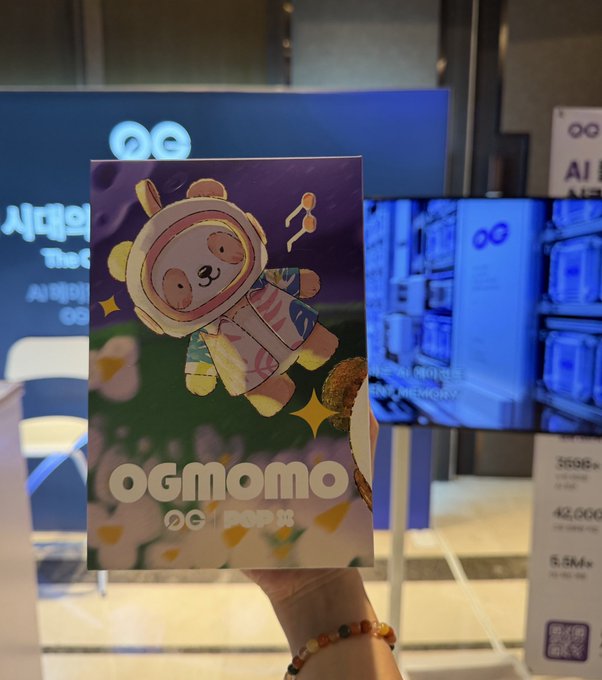 A hand holds a colorful character card labeled 'OGMOMO' from a 'POP MART' x 'OG' collaboration, displayed in front of vending machines.