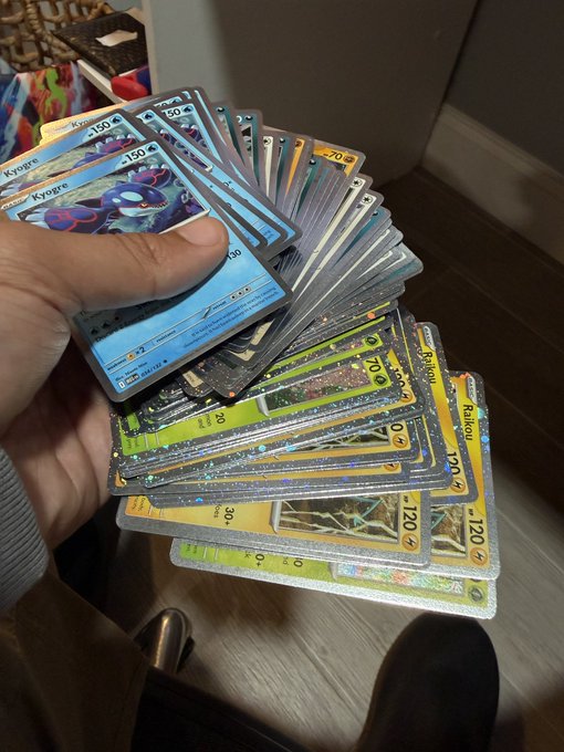 A hand fans out a thick stack of Pokémon trading cards, prominently featuring multiple Kyogre (150 HP) and Raikou (120 HP) holo rares, with higher-HP parallel cards of 70 and 130 visible. The glossy holographic sheen and generous quantity of "Cosmo" cards visually matches the post's excitement about handling a prized collection.