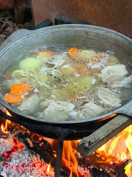 A large pot of soup or stew with carrots and other vegetables is cooking over an open fire, with visible flames and embers beneath it.