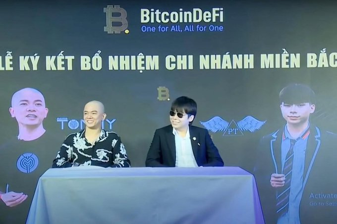 Two men sit at a table during a 'BitcoinDeFi' event, with the banner text reading 'One for All, All for One' and partially visible Vietnamese text indicating a signing ceremony.