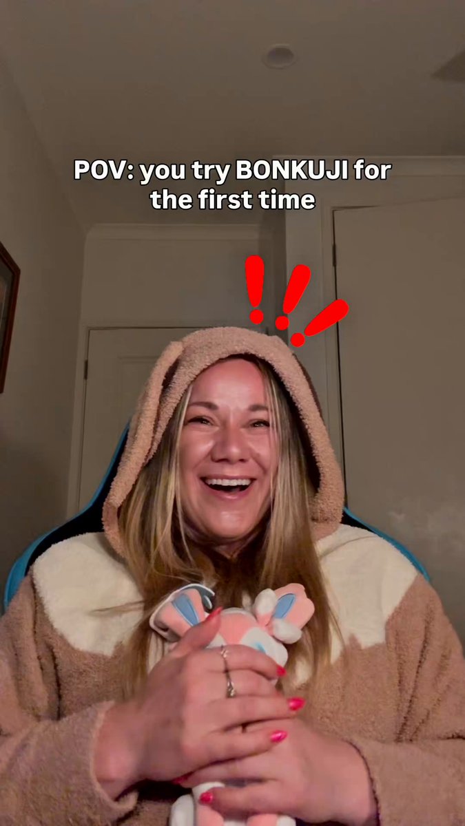 A smiling woman wearing a bear costume, holding a stuffed animal and sitting inside a room with white walls.