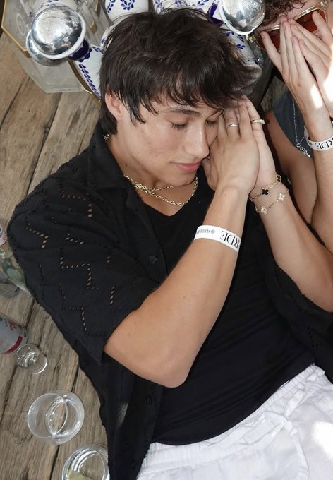 A young man with dark hair wearing a black shirt, gold chain, and luxury bracelets/rings, with his hands pressed together near his face, surrounded by what appear to be ice buckets with champagne bottles.