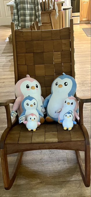 A family of five plush penguin toys in varying sizes and colors (pink, blue, and white) sits on a wooden rocking chair.