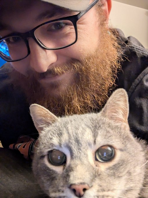 A close-up selfie of a bearded man with glasses posing with a gray tabby cat, both looking at the camera.