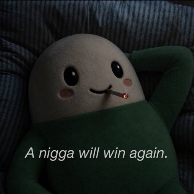 A plush toy character with a smiling face smoking a cigarette, lying in bed under a green blanket, with the caption "A nigga will win again."