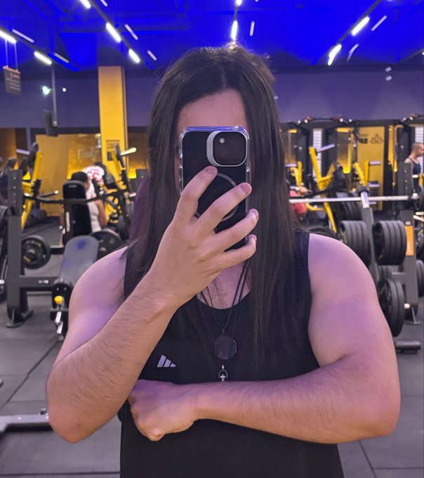 A mirror selfie of a person with long dark hair in a gym, wearing a black Adidas tank top and holding a smartphone in front of their face.