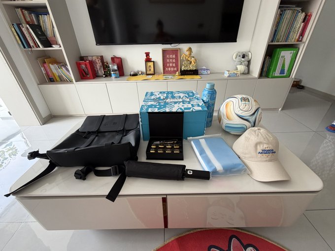 A collection of World Cup-themed merchandise is displayed on a white table, including a black backpack, a beige "Vamos Argentina" cap, a soccer ball, a water bottle, and an open box revealing gold-colored coins or medals. The items are presented as a gift from DeepCoin exchange, complimenting the recipient's World Cup fortune-telling post.