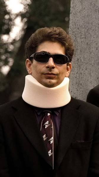 A man with dark hair wearing sunglasses, a suit, purple shirt, striped tie, and a white foam neck brace stands outdoors.