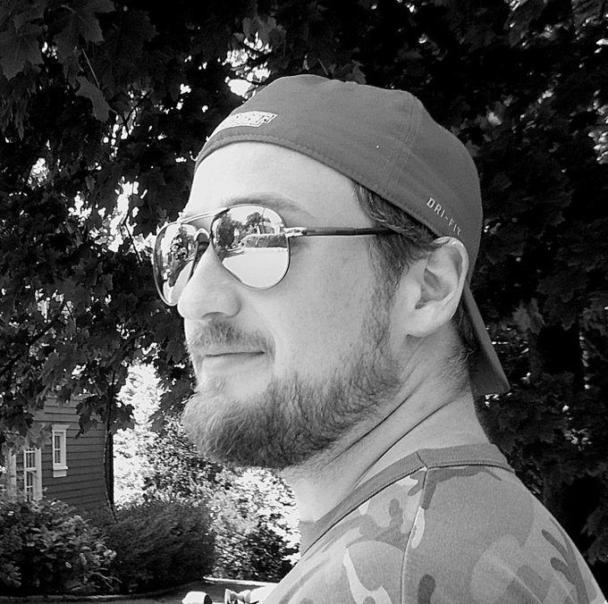 A black-and-white photograph of a bearded man in profile, sporting a backwards baseball cap reading 'DRI-FIT' and reflective aviator sunglasses, with trees in the background.