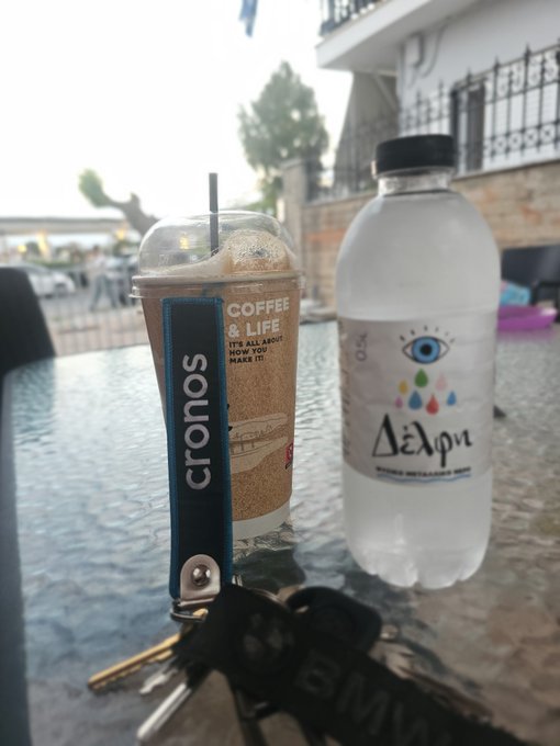 An iced "Cronos" coffee with a sleeve reading "Coffee & Life - It's not a hobby now you make it" sits alongside a bottle of Δέλφι (Delphi) sparkling water on a glass table, with a keychain labeled "BMW" nearby — fueling excitement for a second in-person event.