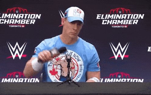 The image features a man wearing a blue shirt, sitting at a table with a microphone in front of him. The background shows a banner that says "ELIMINATION CHAMBER" along with other logos for various companies. The man is holding a microphone and appears to be speaking into it or possibly recording a video.