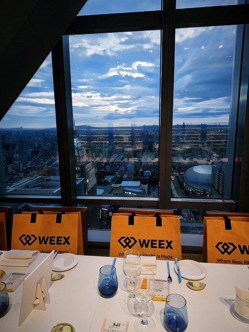 A view from a high-rise restaurant table set with plates, glasses, and yellow shopping bags labeled 'WEEX' with the tagline 'Where Wealth is Made', overlooking a city skyline at dusk.