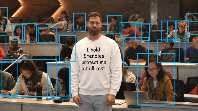 A man wearing a white sweatshirt reading 'I hold $tendies protect me at all cost' stands in a lecture hall, with blue bounding boxes highlighting other students seated around him.
