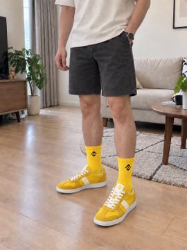 A man in a white t-shirt and dark shorts standing in a living room wearing bright yellow sneakers paired with matching yellow diamond-patterned socks.